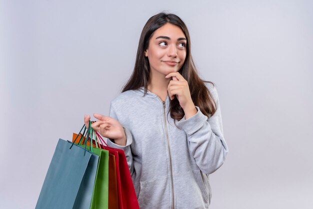 Young beautiful girl in gray hoody looking away with hand on chin thinking pensive holding packages standing over white background