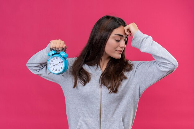 Young beautiful girl in gray hoody looking away with hand on forehead thinking holding clock standing over pink background