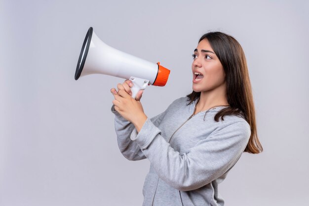 Young beautiful girl in gray hoody looking away with shouting to loudspeaker standing over white background