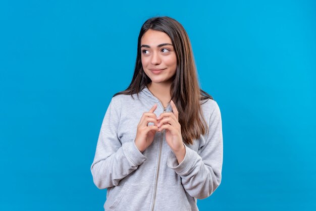 Young beautiful girl in gray hoody looking away with smile on face pleasure standing over blue background