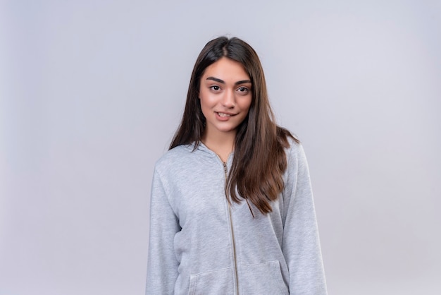 Young beautiful girl in gray hoody looking at camera biting lip standing over white background