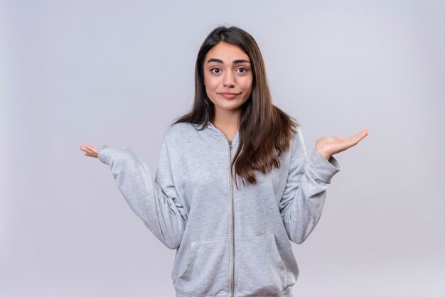 Young beautiful girl in gray hoody looking at  camera pensive on face  having no answer spreading palms standing over white background