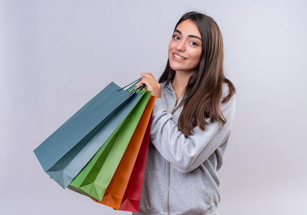 Young beautiful girl in gray hoody looking at camera smile on face with pleasure holding packages standing over white background