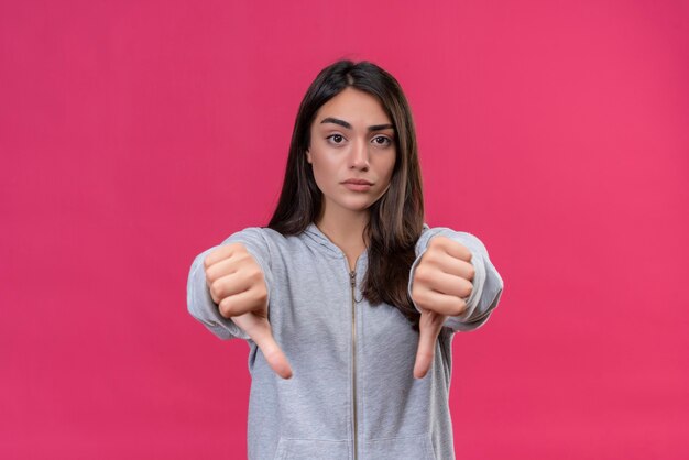 Young beautiful girl in gray hoody looking at camera unpleasant on face and making dislike gesture standing over pink background