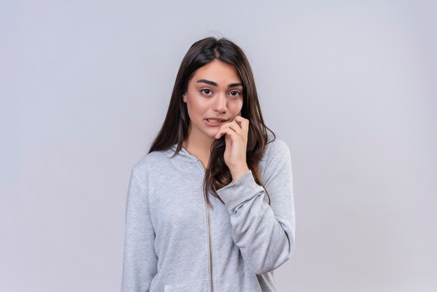 Young beautiful girl in gray hoody looking at camera with confused on face touching cheek standing over white background
