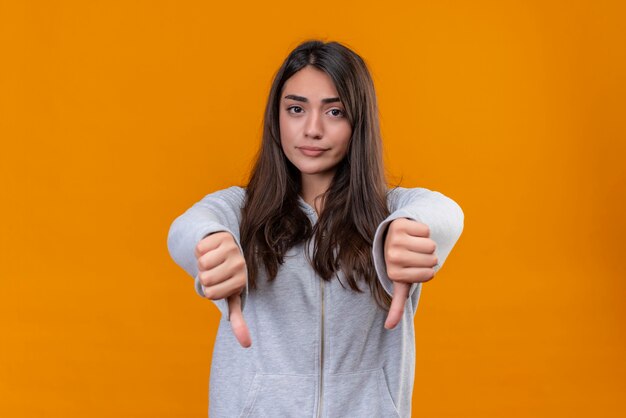 Young beautiful girl in gray hoody looking at camera with dissatisfied sight and making dislike gesture standing over orange background