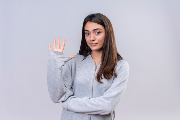 Young beautiful girl in gray hoody looking at camera with pleasant face showing five standing over white background