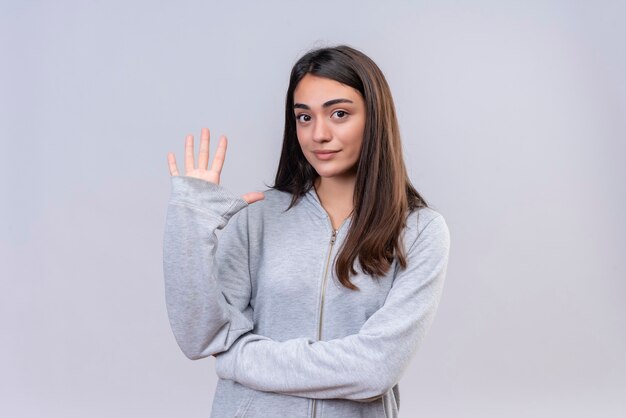白い背景の上に5つの立っているを示す快適な顔でカメラを見て灰色のフーディの美しい少女
