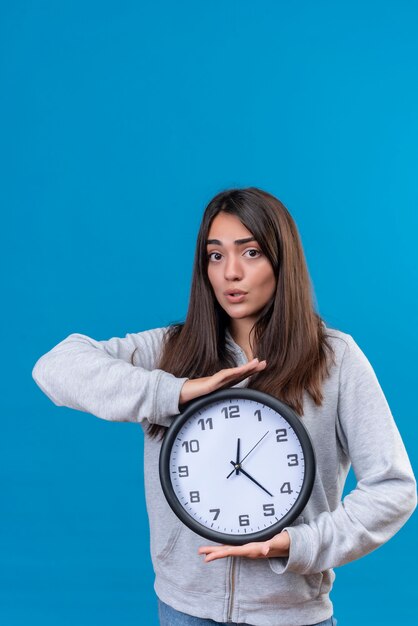 Young beautiful girl in gray hoody looking at camera with shock on face and holding clock standing over blue background