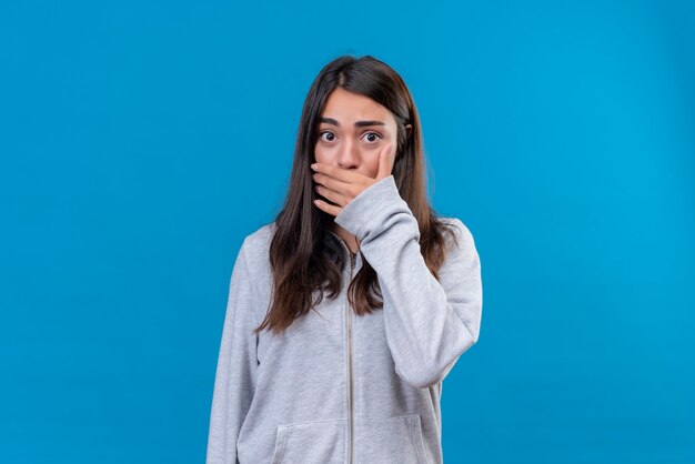 Young beautiful girl in gray hoody looking at camera with shock and hand on mouth standing over blue background