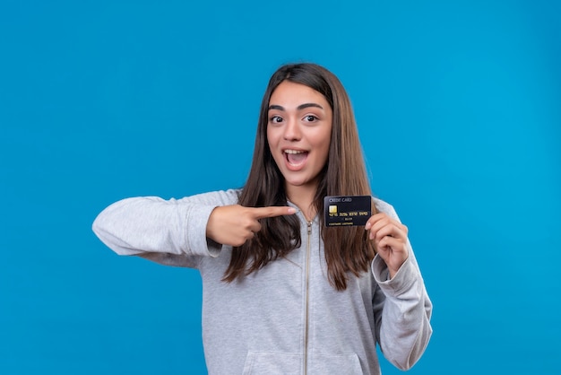 Young beautiful girl in gray hoody  looking at camera with smile on face holding and pointing credit card standing over blue background