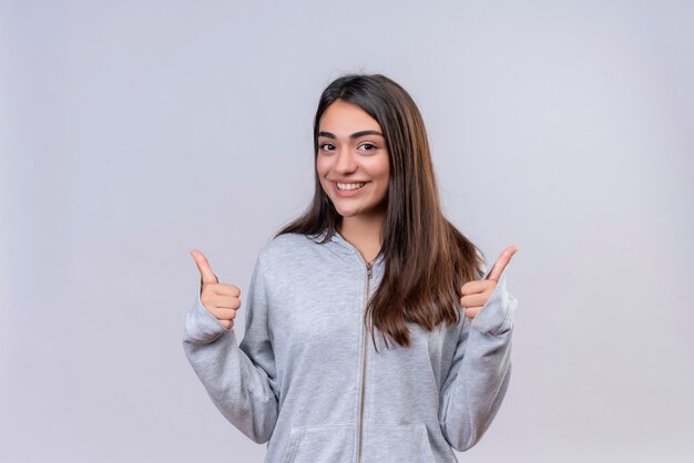 Young beautiful girl in gray hoody looking at camera with smile on face making like gesture standing over white background
