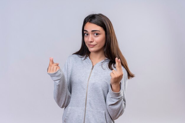 Young beautiful girl in gray hoody looking at camera with smile on face making money gesture standing over white background