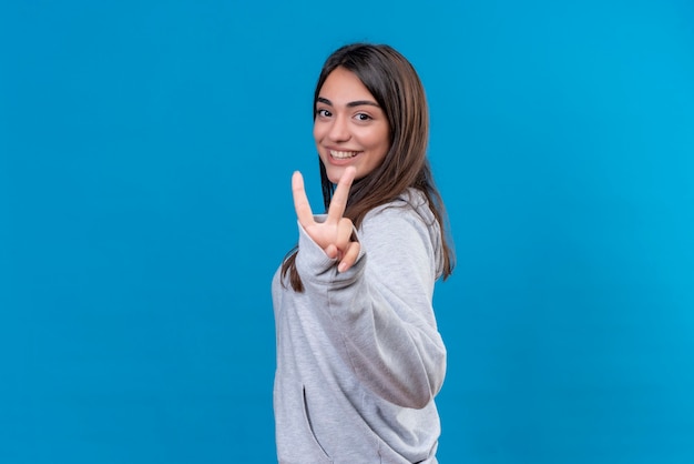 Young beautiful girl in gray hoody looking at camera with smile on face making peace gesture standing over blue background