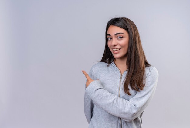 Young beautiful girl in gray hoody looking at camera with smile on face pointing back standing over white background