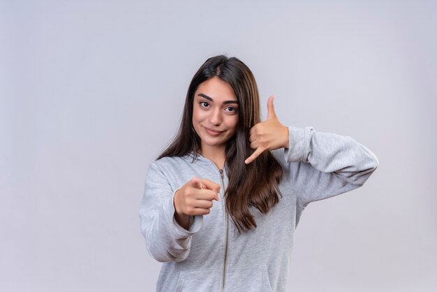 Young beautiful girl in gray hoody looking at camera with smile on face pointing to camera making call me gesture standing over white background