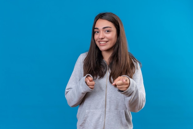 Giovane bella ragazza in felpa con cappuccio grigia che guarda l'obbiettivo con il sorriso sul viso che punta alla fotocamera in piedi su sfondo blu