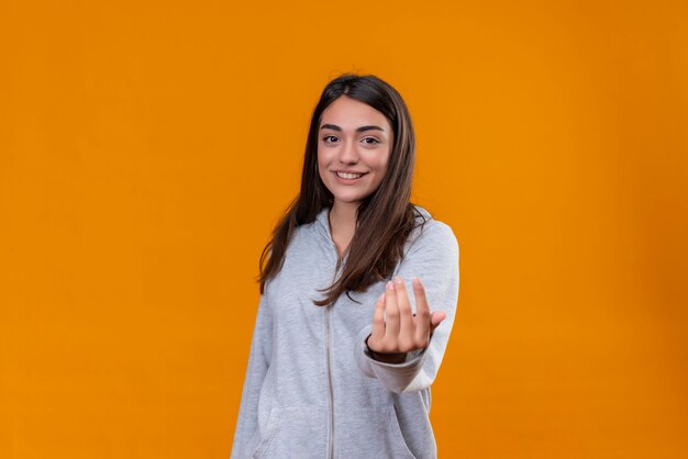 Young beautiful girl in gray hoody looking at camera with smile and reach out standing over orange background