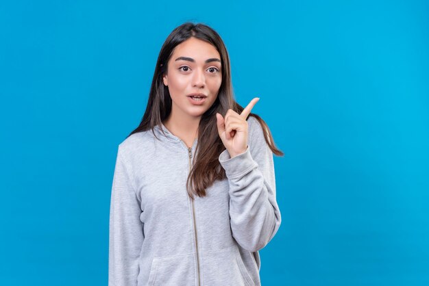 Young beautiful girl in gray hoody looking at camera with surprised sight pointing away standing over blue background