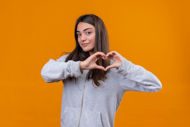 Giovane bella ragazza in felpa con cappuccio grigia che fa gesto romantico del cuore che guarda l'obbiettivo con il sorriso sul viso in piedi su sfondo arancione