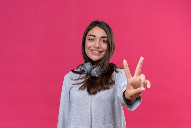 Young beautiful girl in gray hoody wearing headphones looking at camera smile on face making peace gesture standing over pink background