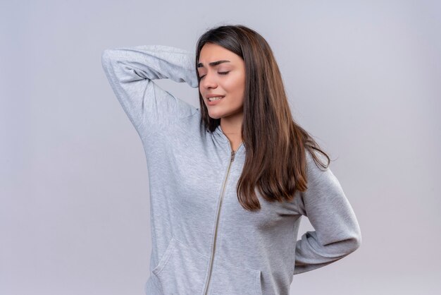 Young beautiful girl in gray hoody with closed eyes touching head for mistake looking confused bad memory concept standing over white background