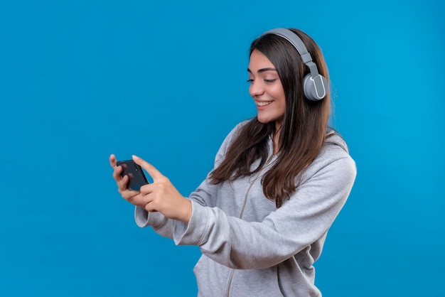 Giovane bella ragazza in felpa con cappuccio grigia con le cuffie che tengono il telefono e guardando il telefono in piedi su sfondo blu