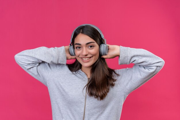 Young beautiful girl in gray wearing headphones  looking at camera with smile on face cheerful touching headphones standing over pink background