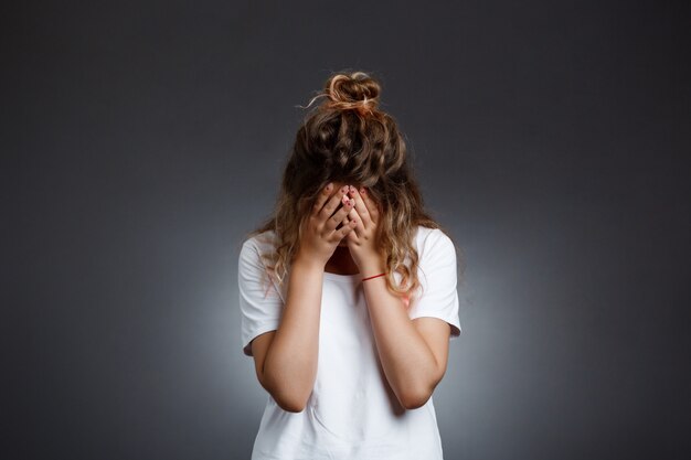Young beautiful girl hiding face with hands over grey wall.