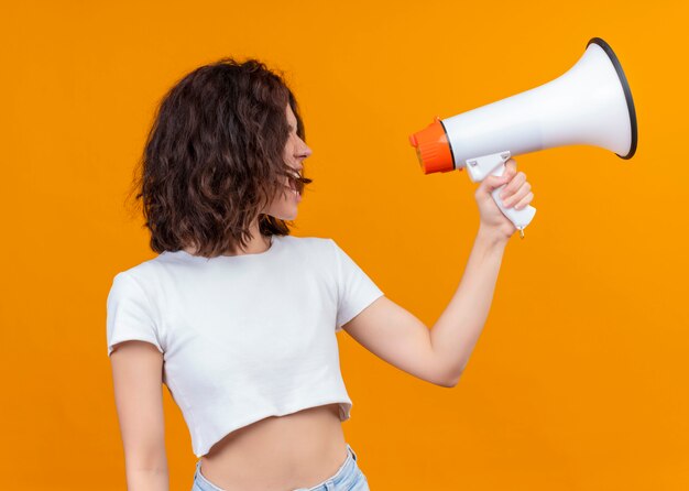 Young beautiful girl holding speaker and talking by it on isolated orange background