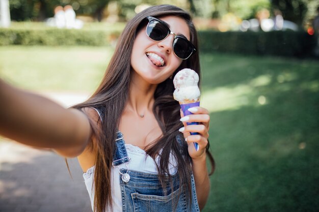 若い美しい少女は白い歯で自分撮り笑顔を作り、サングラスをかけてアイスクリームを保持します