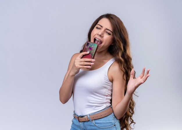 Young beautiful girl singing using mobile phone as microphone with closed eyes on isolated white wall with copy space