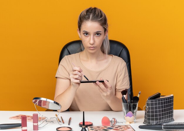若い美しい少女は、オレンジ色の壁に分離された化粧ブラシでアイシャドウを適用する化粧ツールでテーブルに座っています。
