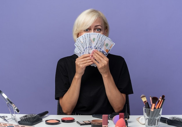 Free photo young beautiful girl sits at table with makeup tools covered face with cash isolated on blue wall