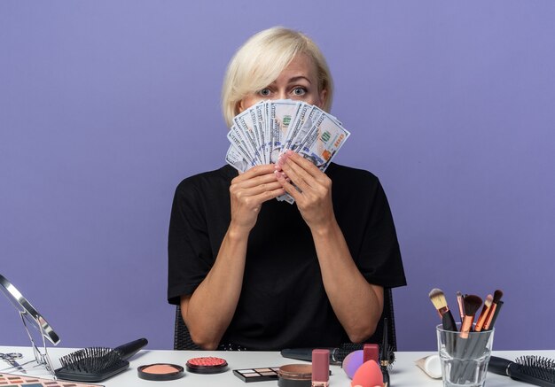 Young beautiful girl sits at table with makeup tools covered face with cash isolated on blue wall