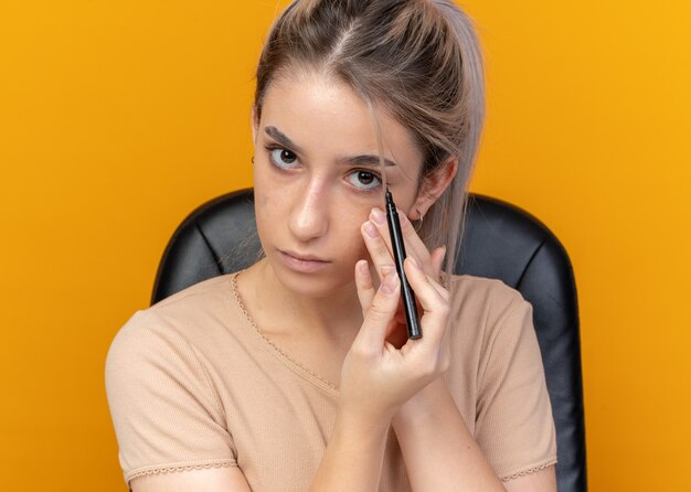  young beautiful girl sits at table with makeup tools draw arrow with eyeliner isolated on orange wall