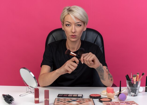  young beautiful girl sits at table with makeup tools holding and crossing powder brushes isolated on pink wall