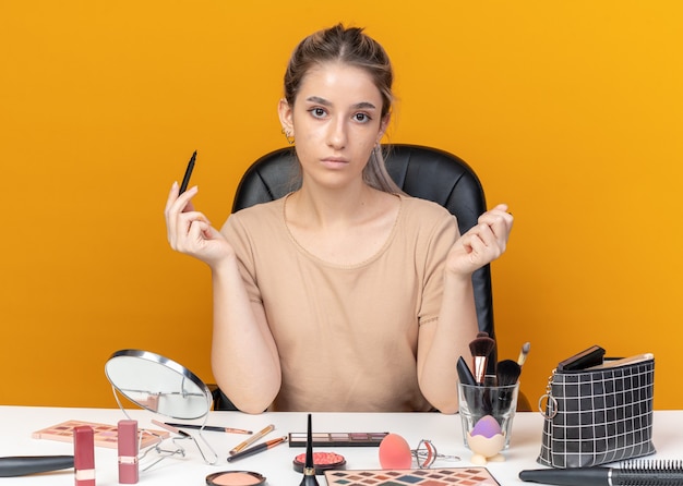  young beautiful girl sits at table with makeup tools holding eyeliner isolated on orange wall