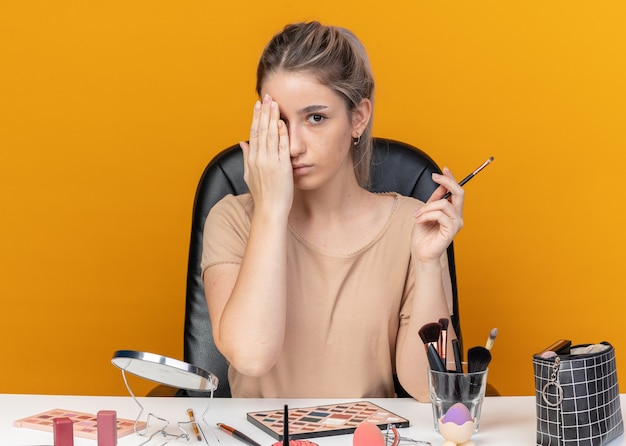  young beautiful girl sits at table with makeup tools holding makeup brush covered eye with hand isolated on orange wall