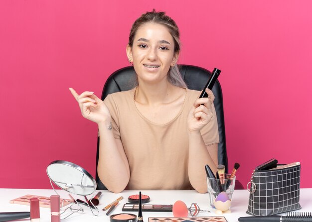 Young beautiful girl sits at table with makeup tools holding powder blush isolated on pink background