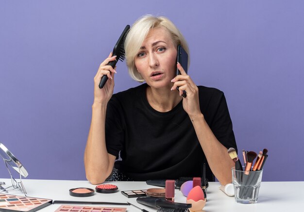  young beautiful girl sits at table with makeup tools speaks on phone combing hair isolated on blue wall