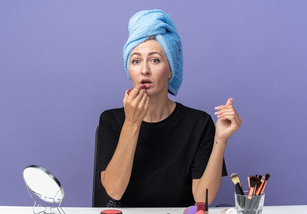 молодая красивая девушка сидит за столом с инструментами для макияжа, вытирая волосы полотенцем, применяя помаду, изолированную на синей стене