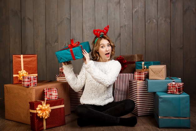 Young beautiful girl sitting among christmas gifts over wooden wall