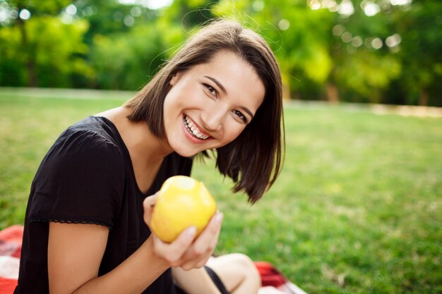 Молодая красивая девушка улыбается, держа яблоко на пикник в парке.