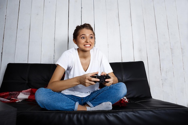 Young beautiful girl smiling, playing video games, sitting on sofa at home. Copy space.