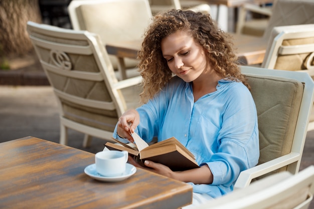 Молодая красивая девушка улыбается, чтение книги, сидя в кафе.