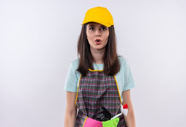 Young beautiful girl wearing apron, cap and rubber gloves looking at camera surprised 