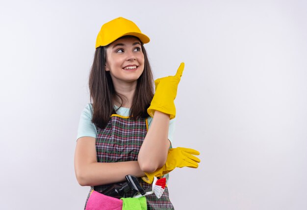 Young beautiful girl wearing apron, cap and rubber gloves smiling cheerfully pointing up with index finger 