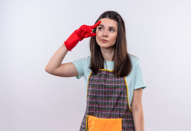 Young beautiful girl wearing apron and rubber gloves looking up pointing her temple trying to remember important thing 