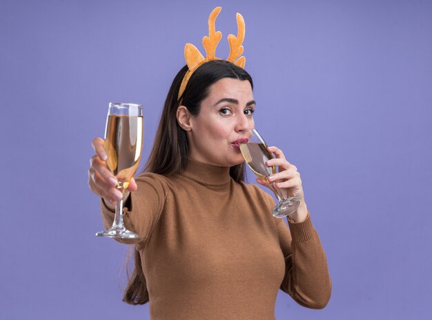 Young beautiful girl wearing brown sweater with christmas hair hoop holding out glass of champagne at camera isolated on blue background